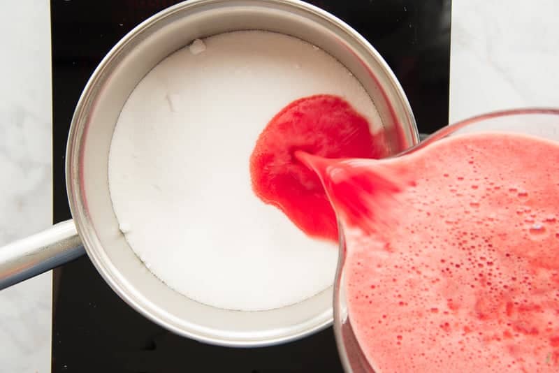 Watermelon juice is poured from a glass bowl into a pot filled with granulated sugar