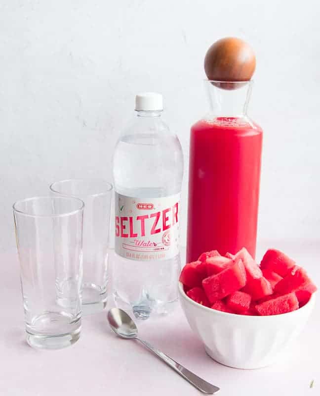 Two highball glasses, a bottle of seltzer water, a pitcher of watermelon simple syrup, and a white bowl filled with cut watermelon