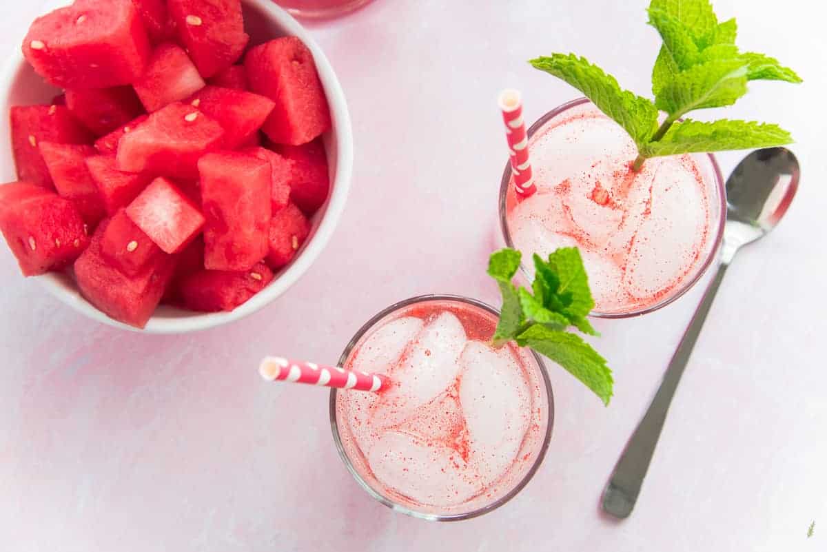 An overhead image of two glasses of Watermelon Italian Sodas with red and white straws a mint stems. A white bowl of watermelon sits to the left