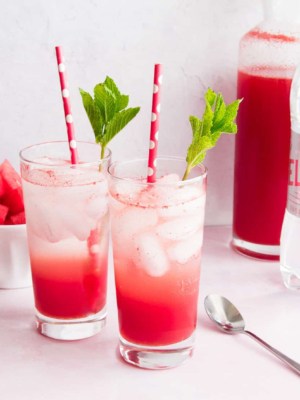 Two tall glasses filled with Watermelon Italian Soda. Mint leaves and red and white straws in each glass. A bowl of watermelon and two bottles in background