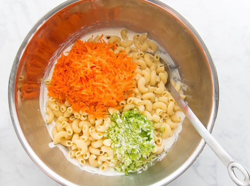 Cooked macaroni pasta, chopped green onions, shredded carrots, are added to the dressing in a large metal bowl. A metal spoon is on the right side of the bowl ready to mix the salad together.