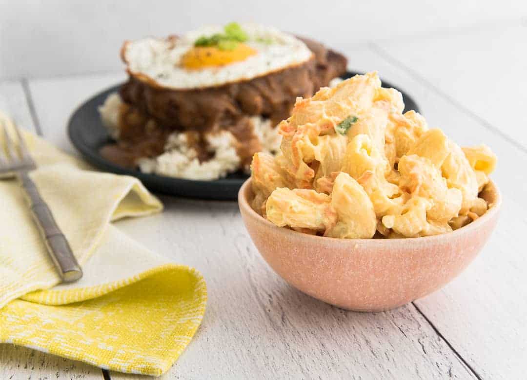 A small pink bowl is filled with Macaroni Salad. To the left of the bowl is a furled yellow napkin and a silver fork. In the background is a blue plate with rice and loco moco
