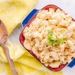 Overhead shot of blue dish filled with Macaroni Salad on right of image. Left is a furled yellow napkin on which lays a wooden spoon