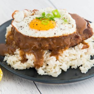 A close up image of a blue plate with white rice, loco moco, and a fried egg on it. A corner of a yellow napkin is in the left corner and fork peeks out from upper left corner