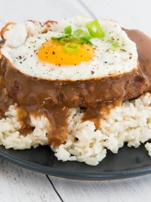 A close up image of a blue plate with white rice, loco moco, and a fried egg on it. A corner of a yellow napkin is in the left corner and fork peeks out from upper left corner