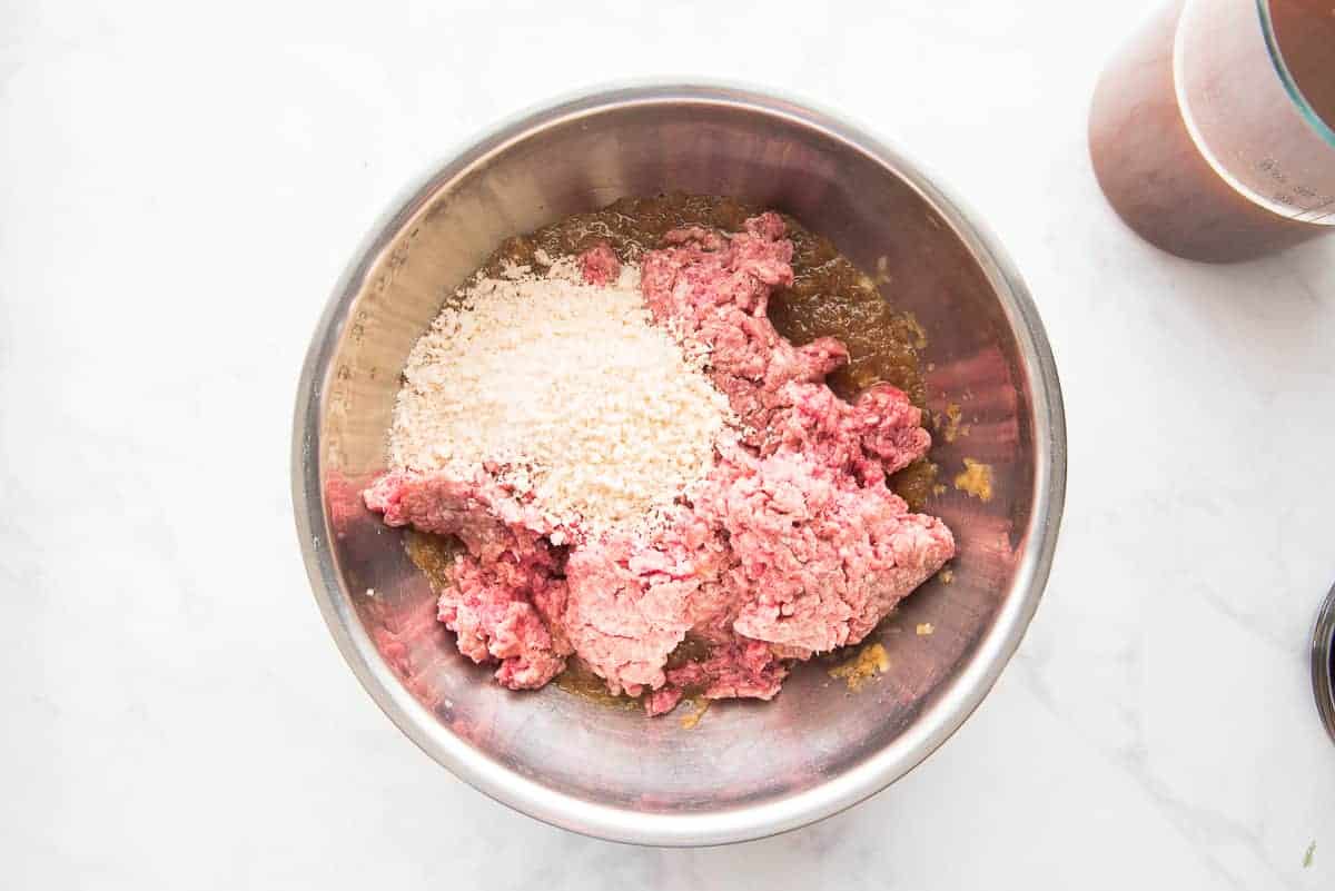 Breadcrumbs and ground beef are add to the onion paste in a silver mixing bowl