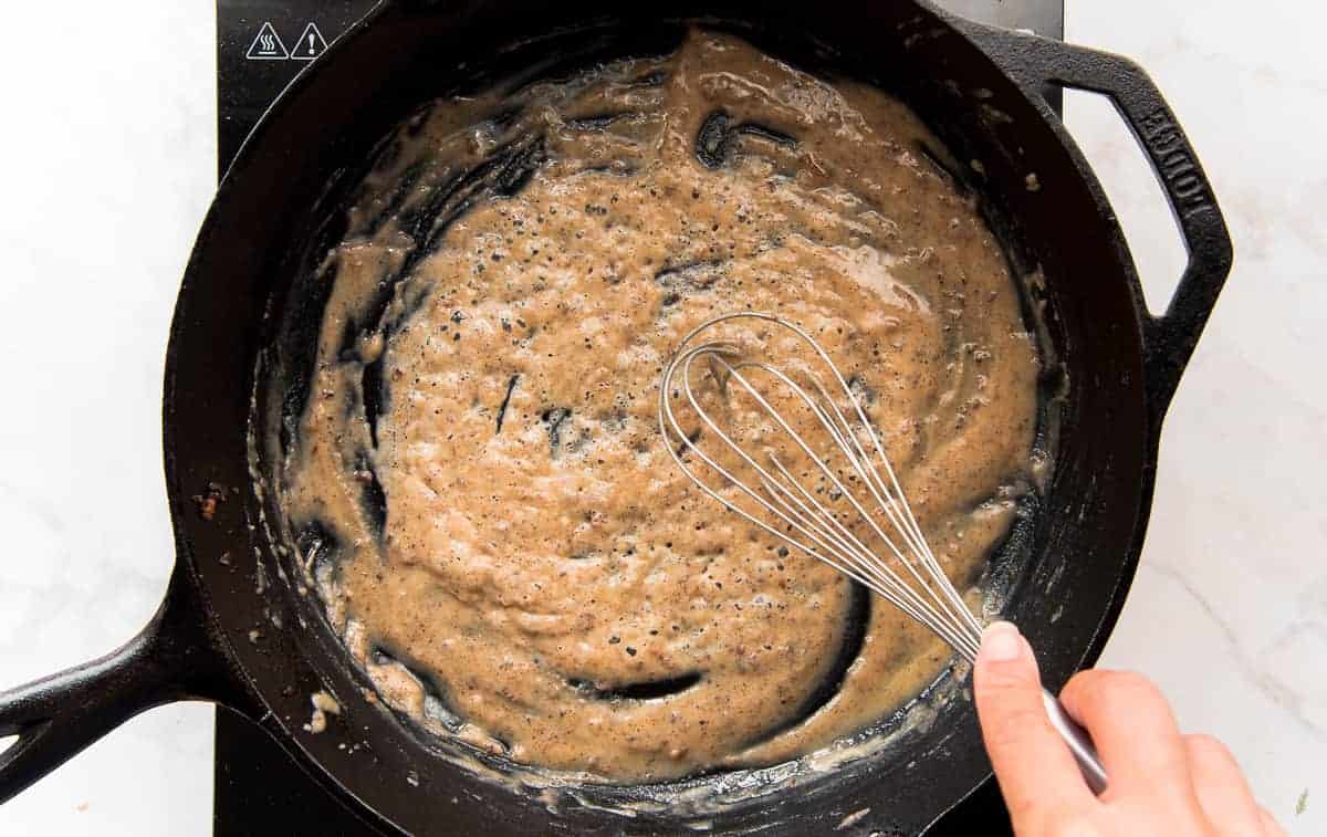 A whisk is used to stir the brown roux in a cast iron skillet