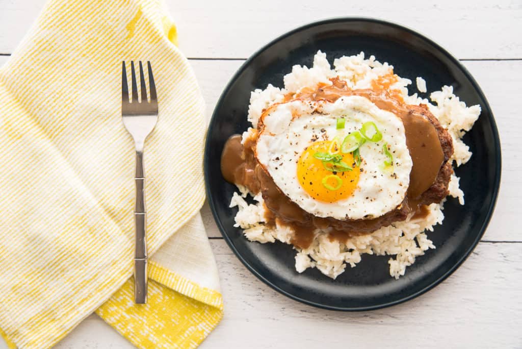 A plate of loco moco, consisting of a hamburger patty topped with a sunny-side-up egg and brown gravy, served on a bed of white rice. A fork and a yellow-striped napkin are placed next to the plate on a white surface.