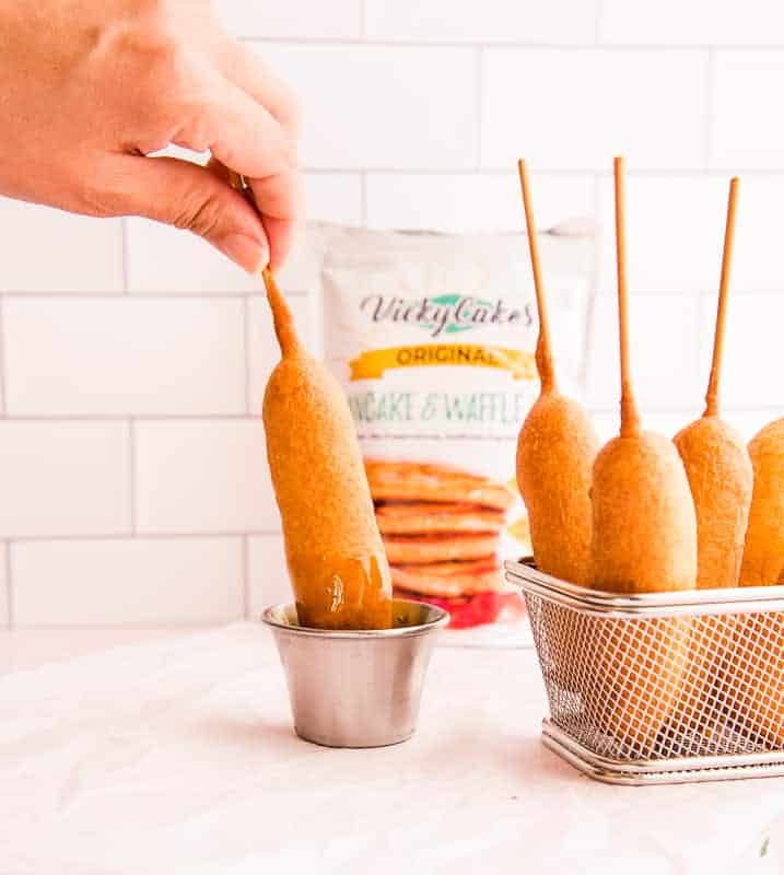 A hand dips a stick of Pancakes and Sausage on a stick into a silver cupful of syrup. A basket of Pancakes and sausage on a stick is on the right. A white pouch of pancake mix is in the background.