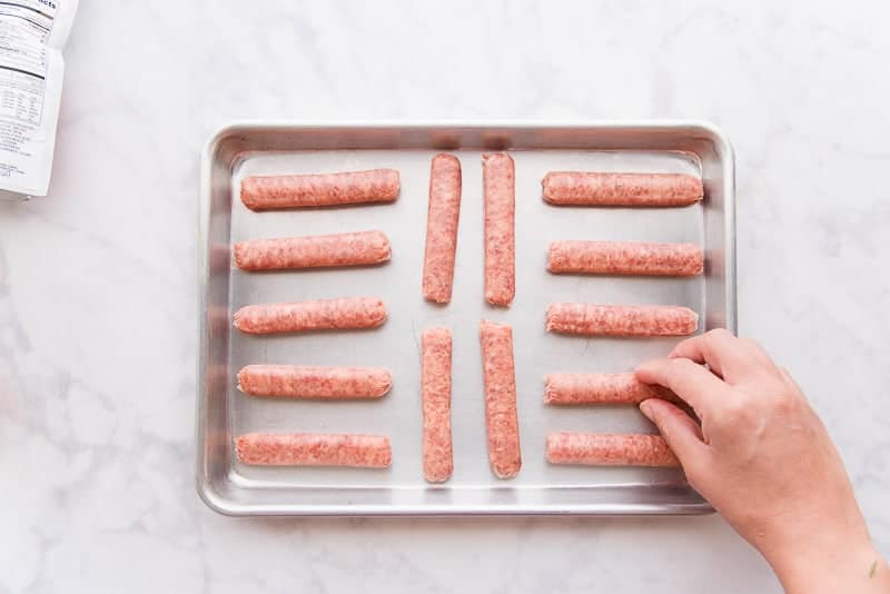 A hand arranges breakfast links on a silver sheet pan