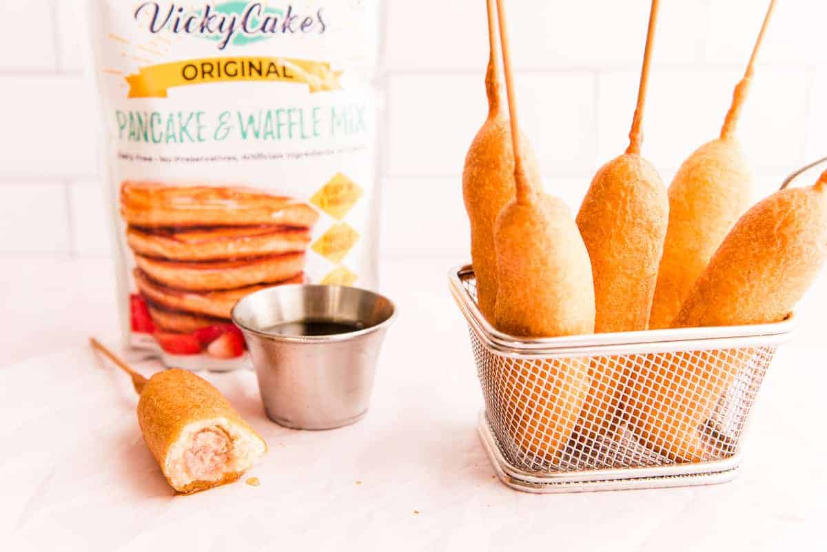 A landscape image of a basket filled with Pancakes and Sausage on a stick. A white pouch of pancake mix is in left background. A stick with a bite taken out is left of the basket next to a silver cup filled with syrup