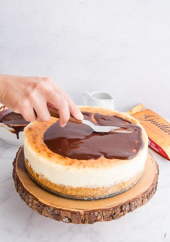 A hand spreads chocolate ganache over the surface of the toasted marshmallow cheesecake. A gold bag of chocolate chips is in the right back corner.