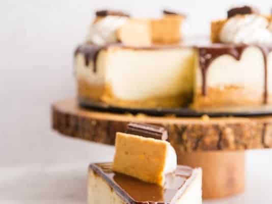 A fork has a bite of S'mores Cheesecake on it. It sits on a black plate in front of a wooden cake stand with the rest of the cheesecake on it.