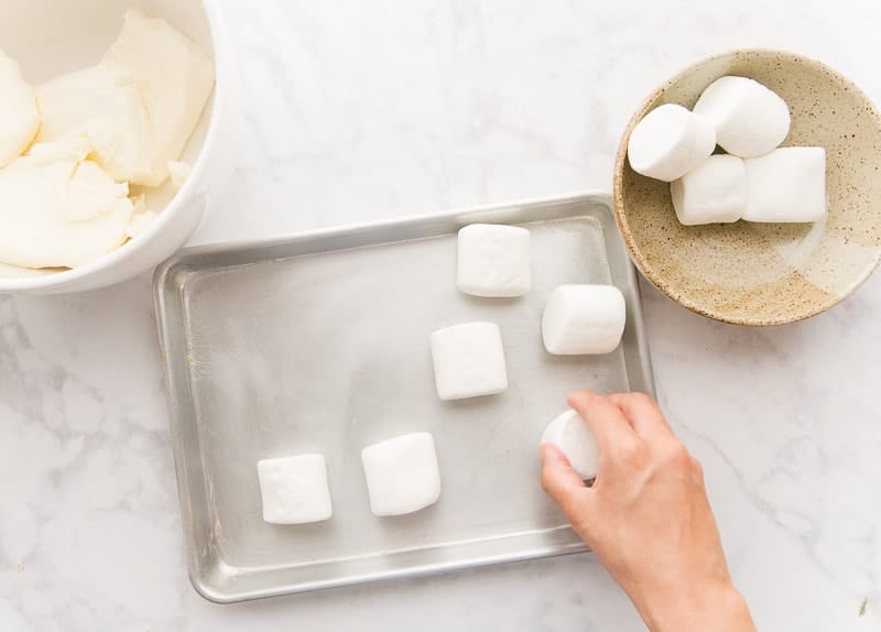 A hand arranges jumbo marshmallows onto a lightly greased sheetpan. A brown bowl filled with marshmallows is in the top right corner. A white bowl with cream cheese is in top left.