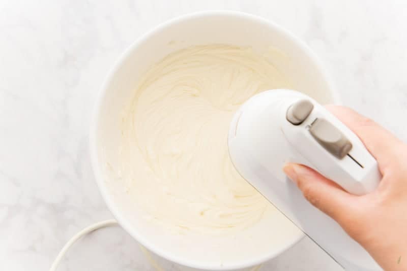 An electric hand mixer beat the cream cheese, sugar, and eggs together in a white ceramic bowl.