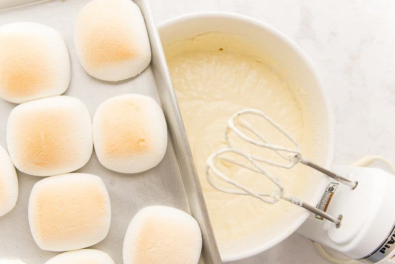 Toasted marshmallows are held on a sheet pan over the cheesecake batter which is in a white bowl. An electric hand mixer is in the bottom right hand corner of the image