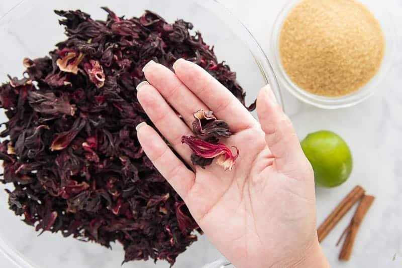 A hand holds dried hibiscus flowers above a bowl filled with more flowers. Next to the bowl are limes, cinnamon sticks, and a bowl of raw cane sugar.