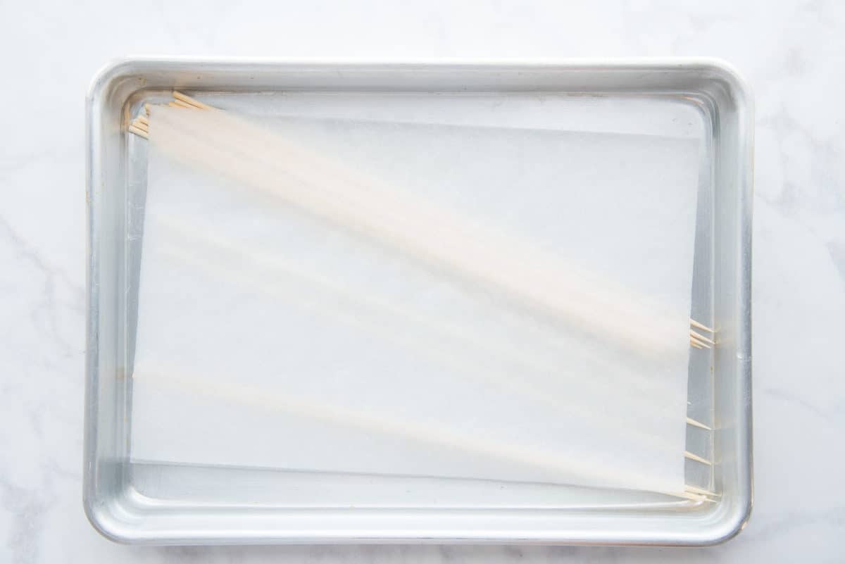 Skewers are submerged in water under a white paper towel on a sheetpan