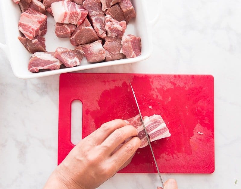 Hands use a knife to cut pieces of pork smaller on a red cutting board.