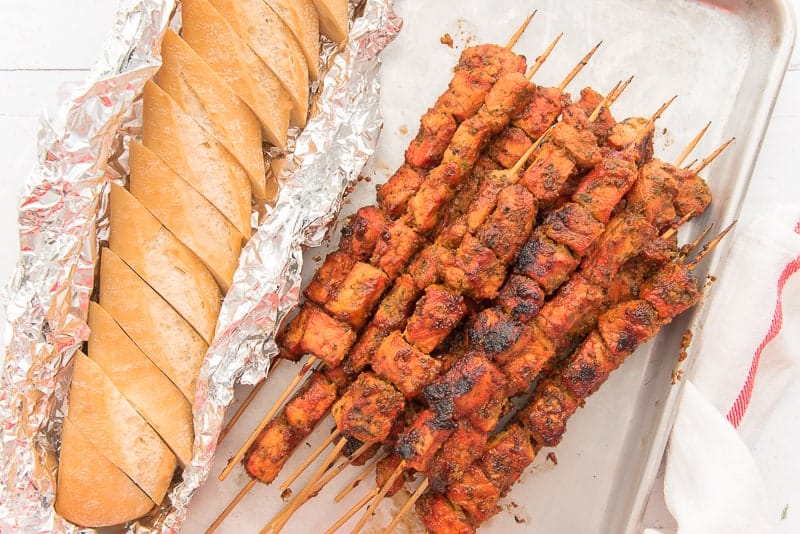 A close-up horizontal image of the Pinchos on a sheetpan next to a loaf of bread sliced in aluminum foil