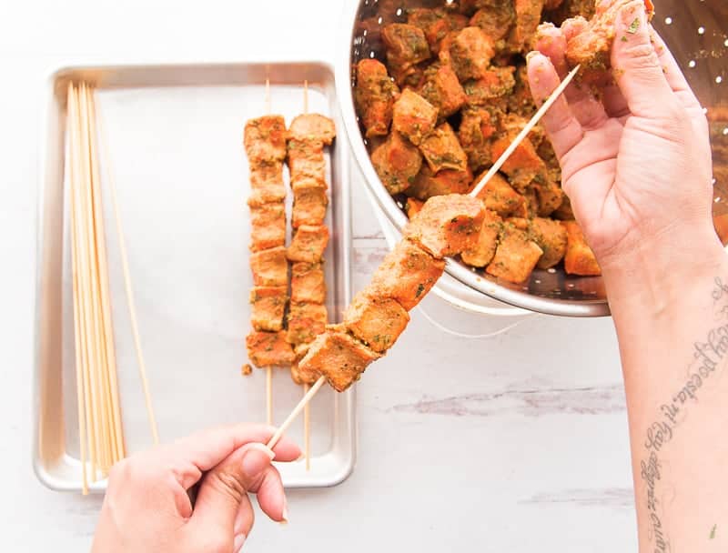 A person skewering the marinated pork cubes onto wooden skewers. More skewers are on a metal sheetpan.