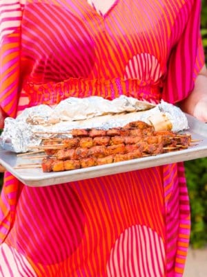 A person in a brightly colored caftan holds a sheetpan of Pinchos in front of greenery.
