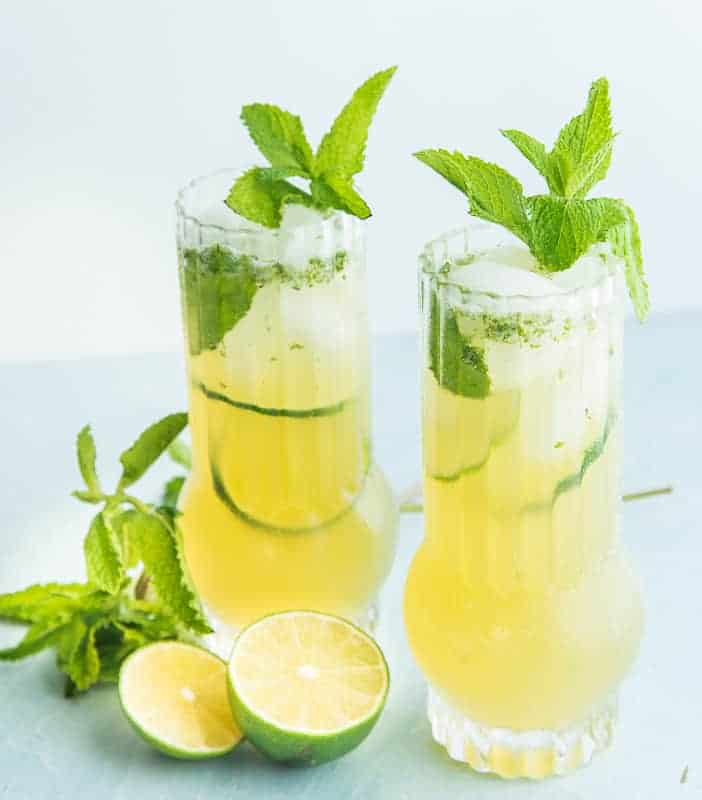 Two highballs of Puerto Rican Mojitos, sliced limes and mint sprigs lie around the glasses