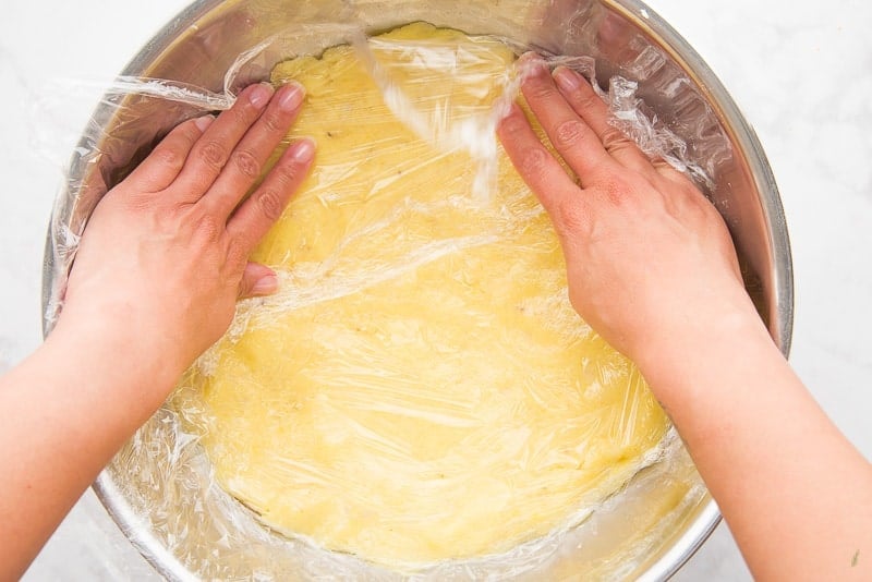 A hand presses plastic wrap onto the mashed potato mixtures in a silver bowl for the rellenos de papa