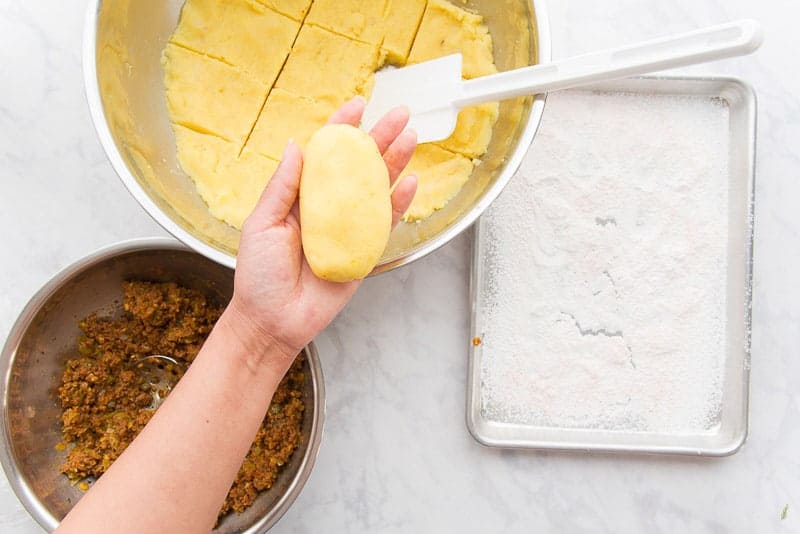A hand holds a formed relleno de papa above a bowl filled with mashed potatoes and another filled with picadillo. A metal pan is covered in cornstarch