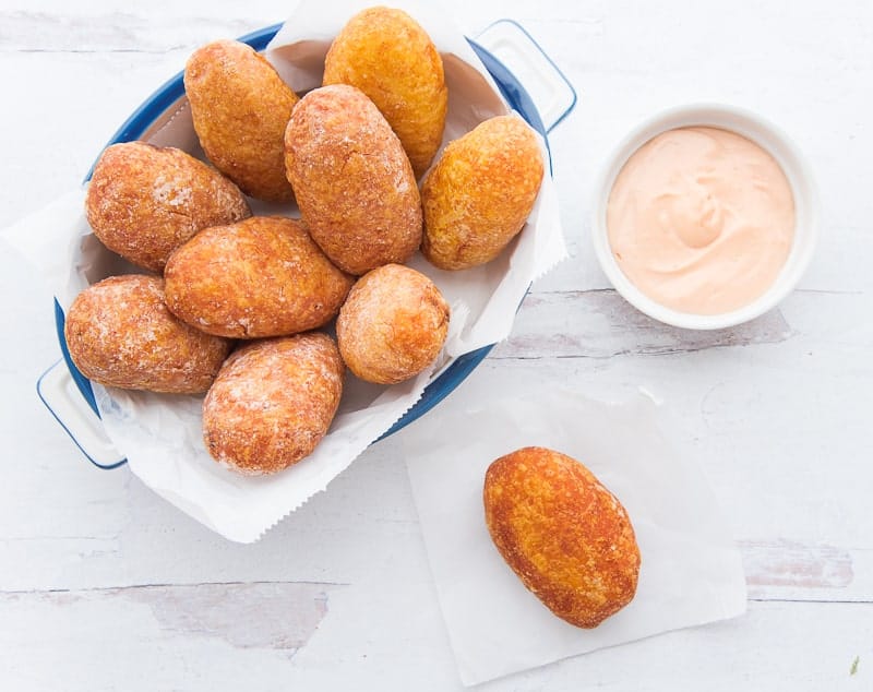 White, blue-rimmed pan is filled with rellenos de papa. A relleno is on a square of wax paper below a white bowl filled with mayo-ketchu sauce.