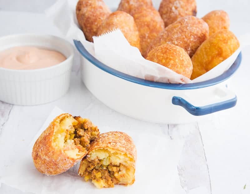 A relleno de papa is broken open to reveal the interior meat filling. A white pan with a blue rim and handles is to the right. A white dish filled with pink sauce is on left