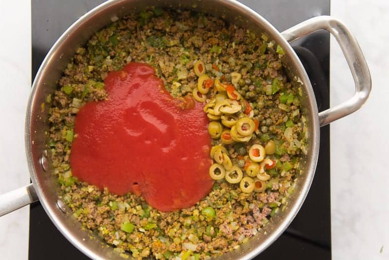 Tomato sauce, sliced olives, and capers are added to the beef picadillo in a silver pan