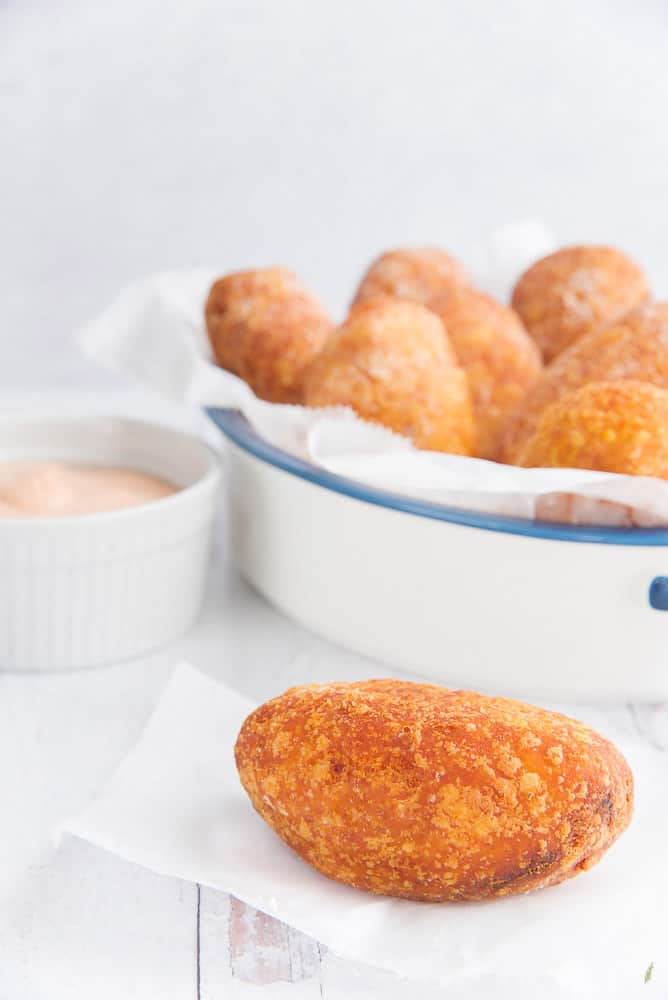 A single relleno de papa is on a white piece of wax paper next to a white bowl filled with pink sauce Rellenos de papa in a white pan with blue trim.