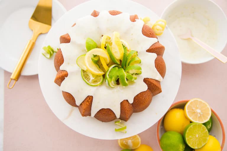 A beautifully decorated bundt cake topped with white icing and garnished with lemon and lime slices. It sits on a white plate with a gold cake server nearby. A bowl of fresh lemons and limes is in the background.
