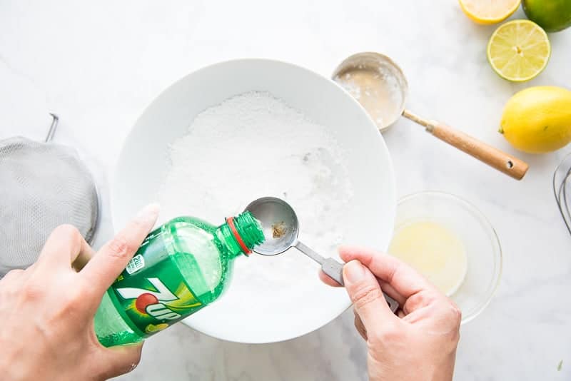A hand measures a spoonful of 7up into a measuring spoon which is held over a white bowl filled with powdered sugar
