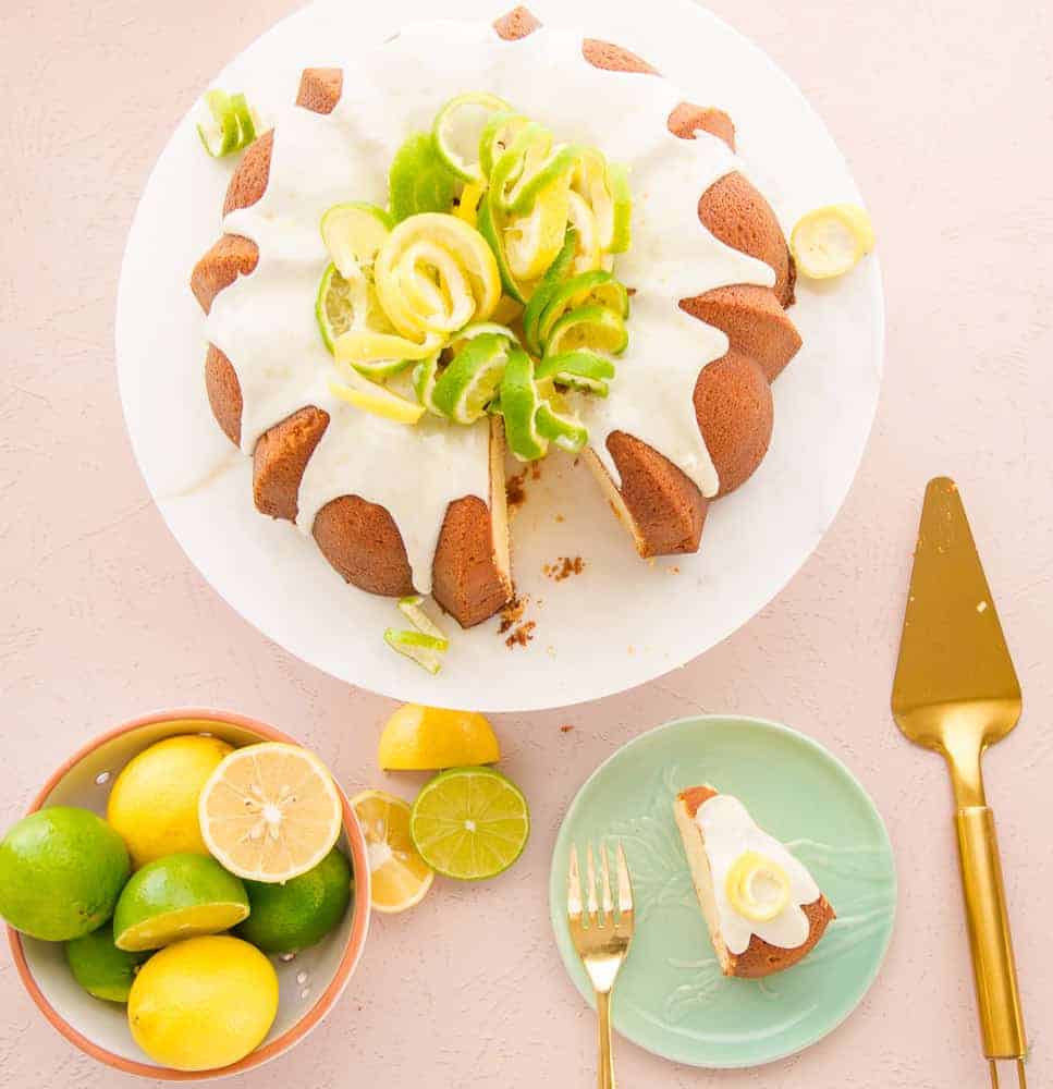 An overhead image of the poundcake glazed and decorated with citrus peels. A slice of cake is removed and sits on a green plate with a gold cake server on the right. A white bowl of citrus is on the left