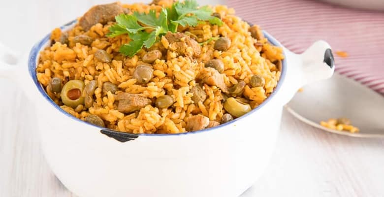 A white, blue-rimmed bowl filled with Arroz con Gandules, garnished with sprigs of fresh cilantroa silver spoon in background has grains of rice on it.