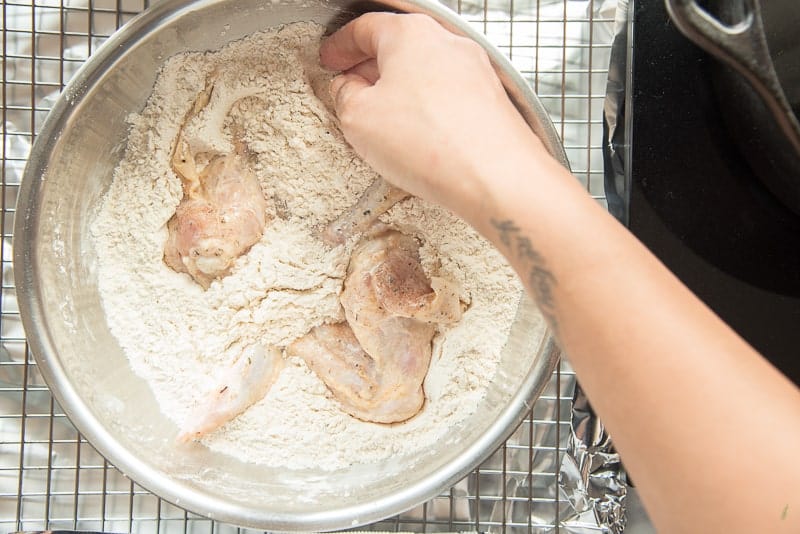 A hand breads the marinated chicken in seasoned flour in a silver bowl over a wire rack.