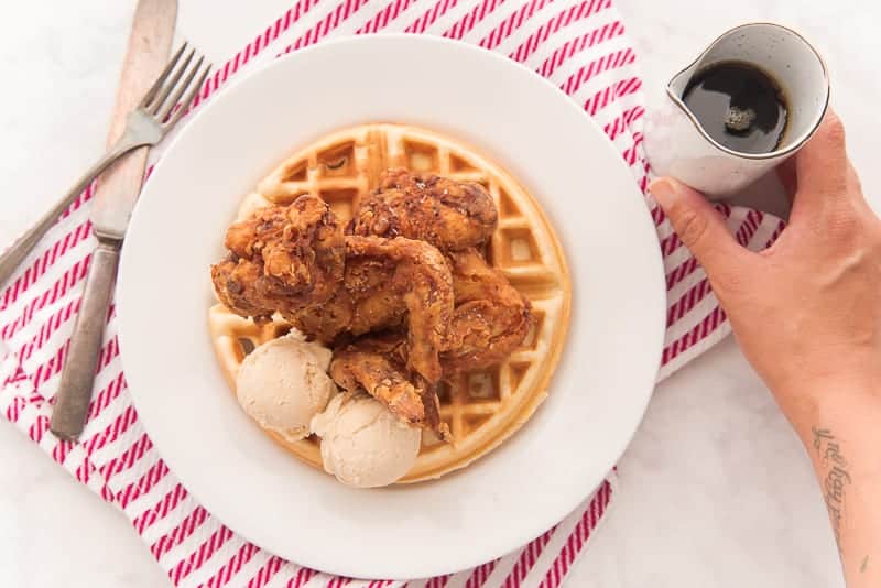 A hand grabs a white pitcher of maple syrup to the left is a white plate with Chicken and Waffles over a red and white striped towel. A knife and fork are crossed to the left of the plate