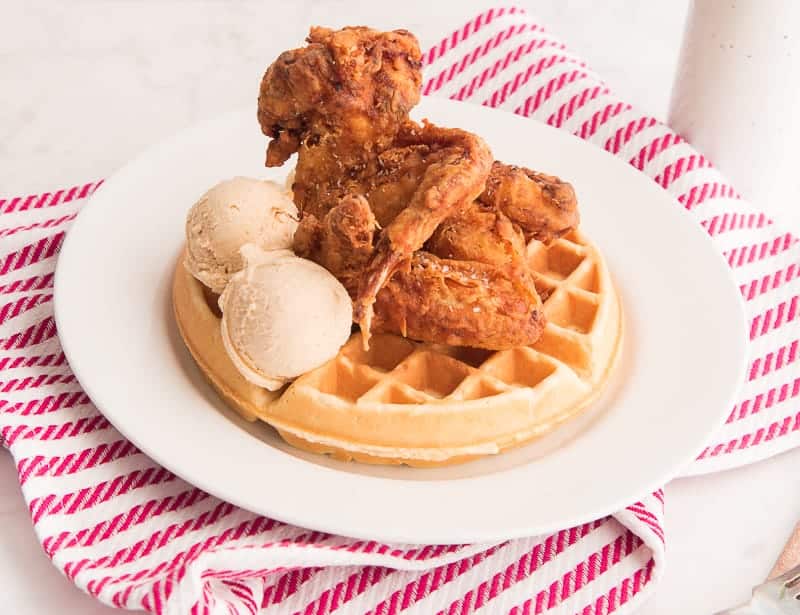 A white plate over a red and white striped towel on the plate are Chicken and Waffles with two scoops of Maple-Cinnamon Butter