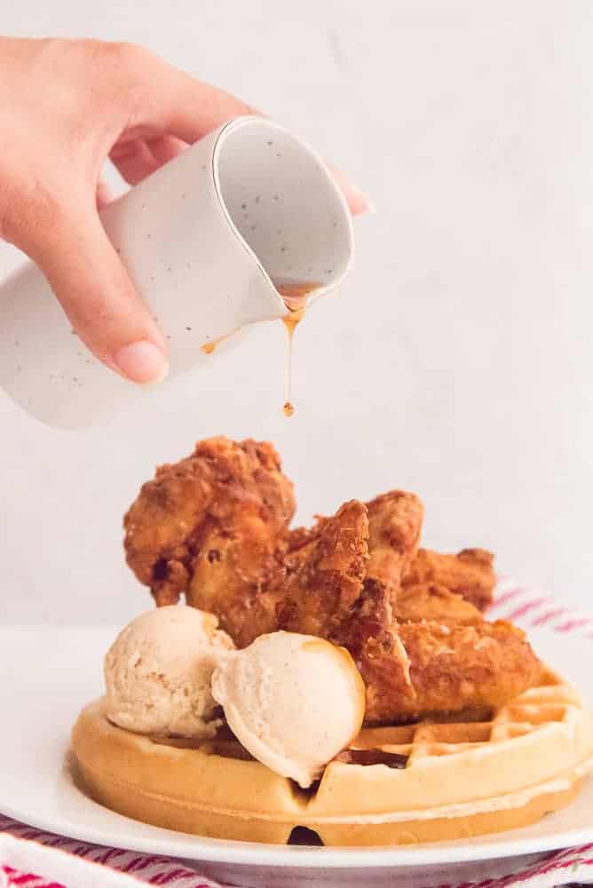 A hand pours syrup over chicken and waffles on a white plate