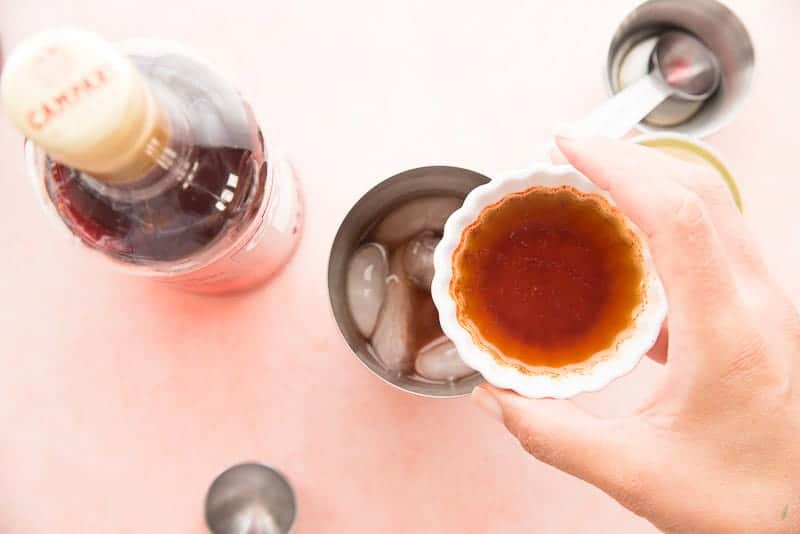A hand holds a bowl of chipotle simple syrup over the cocktail shaker filled with the rest of the cocktail ingredients