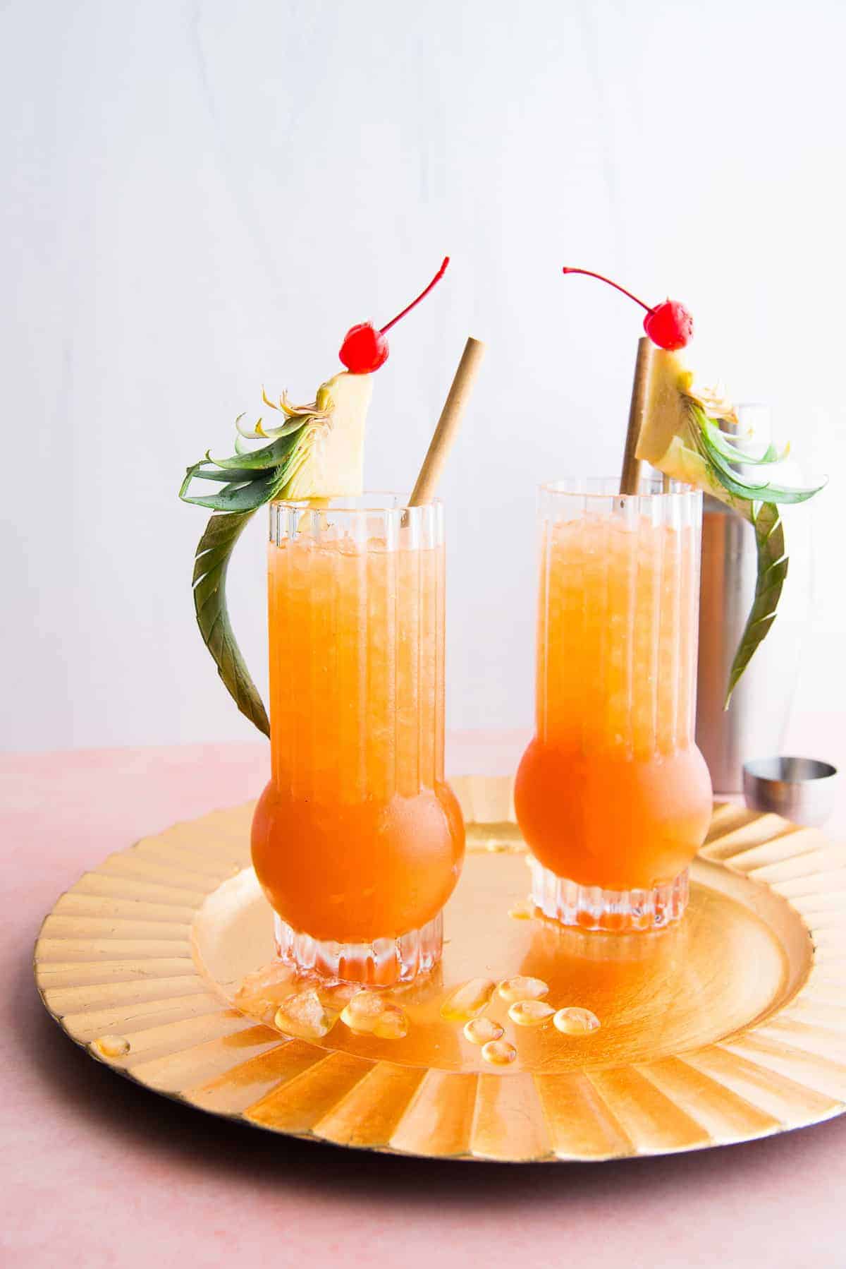 Two Jungle Bird cocktails in highball glasses on a gold tray on a coral top with a blue background