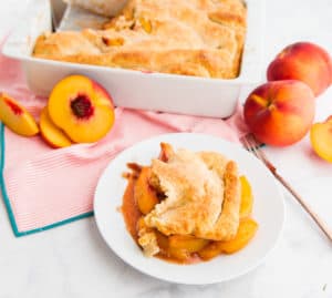 A serving of Old-fashioned Peach Cobbler on a white plate. The baking dish of peach cobbler is in the background next to peaches.