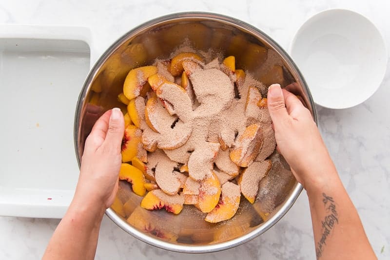 Hands toss the sliced peaches in the brown-sugar cornstarch mixture in a silver bowl.