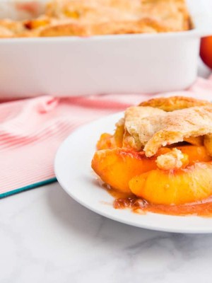 A serving of Old-fashioned Peach Cobbler on a white plate. Peach halves are in the background