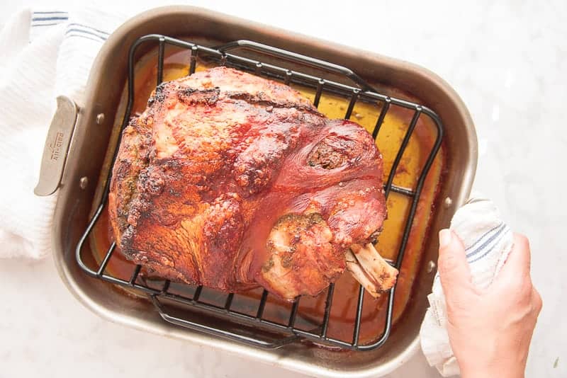 The finished pernil is in a roasting pan. A hand grips the pan with a white and blue towel