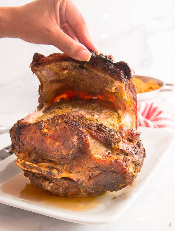 A hand lifts the chicharron from the pernil which is on a white platter