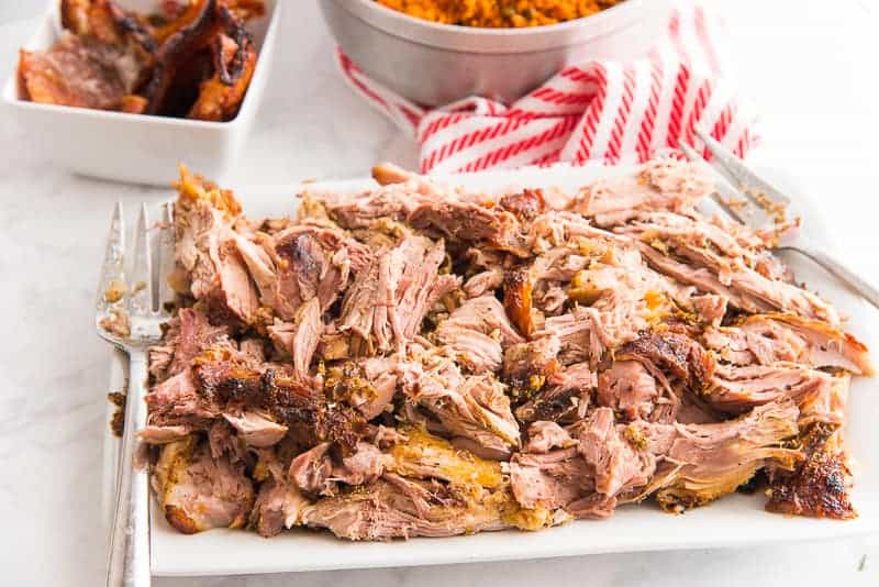 A horizontal image of a white platter of shredded Pernil. A furled orange and white striped towel is behind.