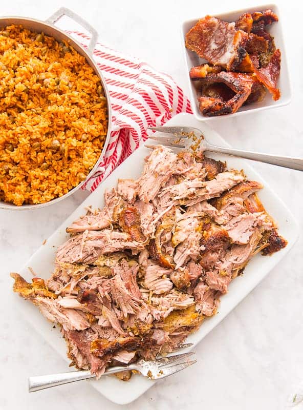 Shredded pork is on a white platter with two serving forks on either end. A silver pot filled with rice and pigeon peas top left. A white bowl with pork crackling top right.